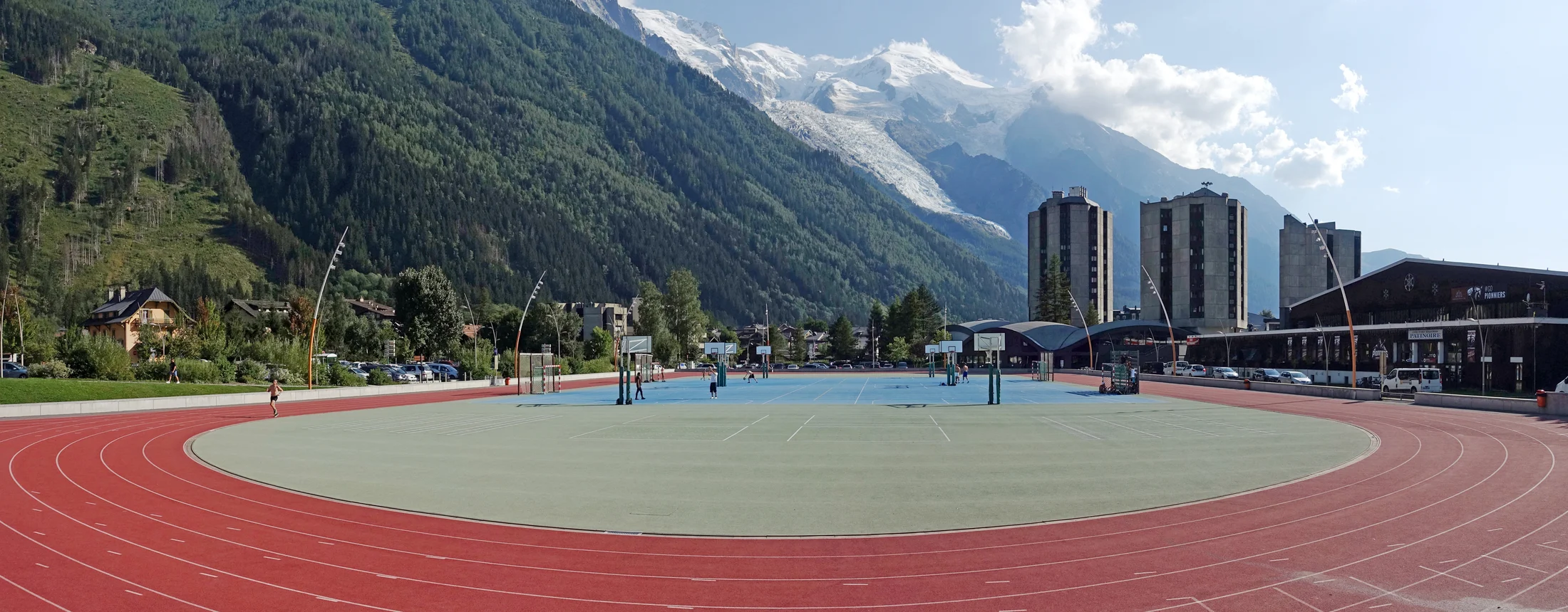 Red running lanes curving around the Chamonix multisports courts, with forested slopes and glaciated mountains above the town.