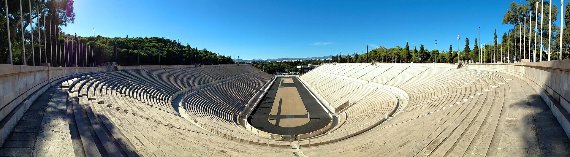 A panoramic view of the white marble seating and narrow running surface of the Panathenaic Stadium in Athens beneath a blue sky.