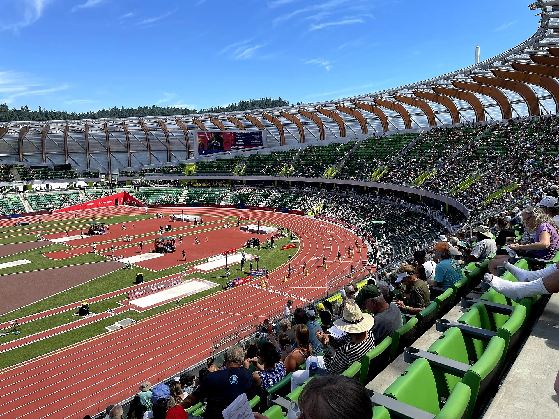 The red track at Hayward Field curving below sweeping timber roof supports, with spectators in green seats at the 2021 US Olympic Trials.