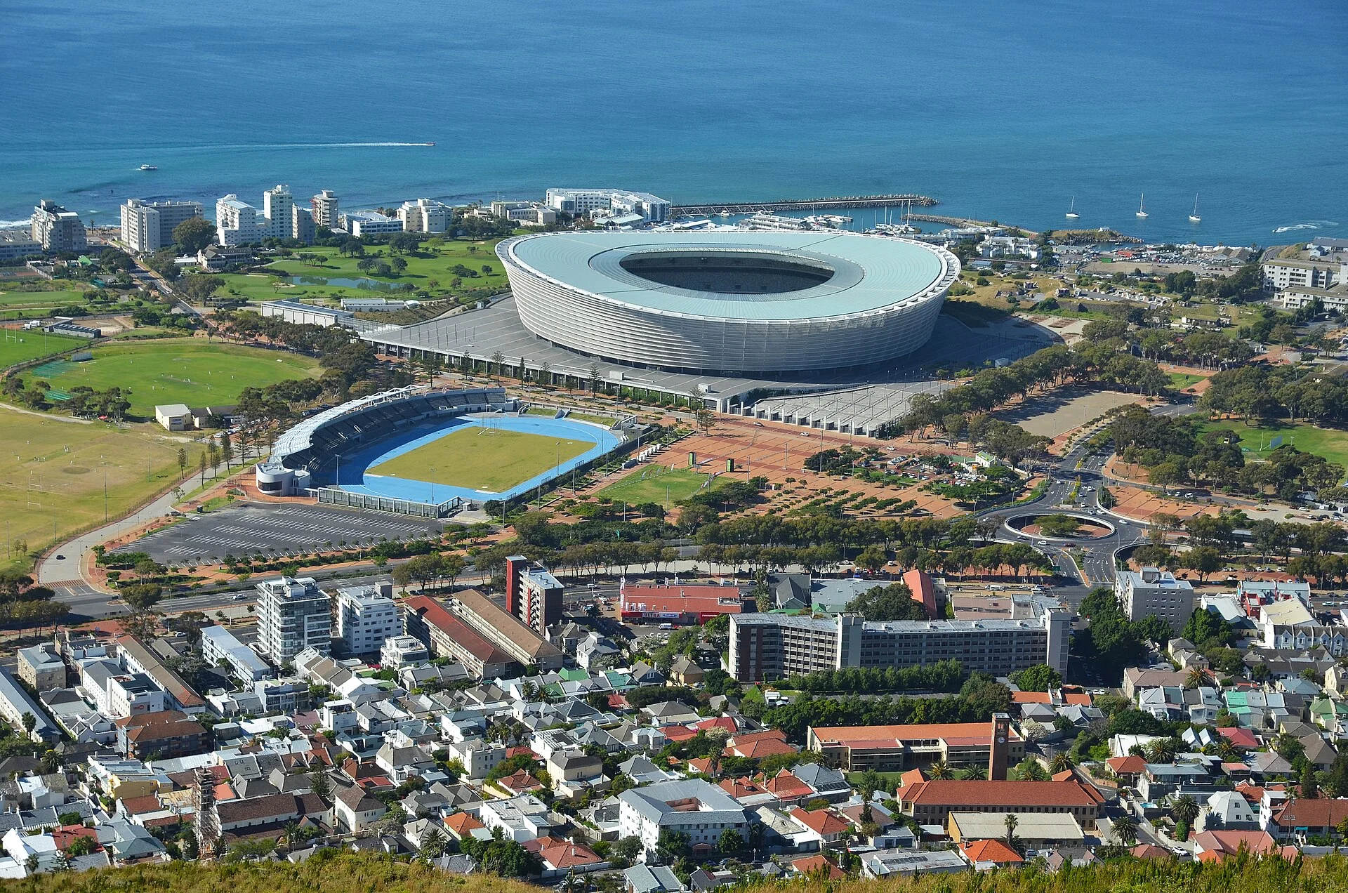 Aerial view of the blue athletics oval at Green Point beside Cape Town’s large white football stadium, with neighbourhoods and the Atlantic Ocean beyond.