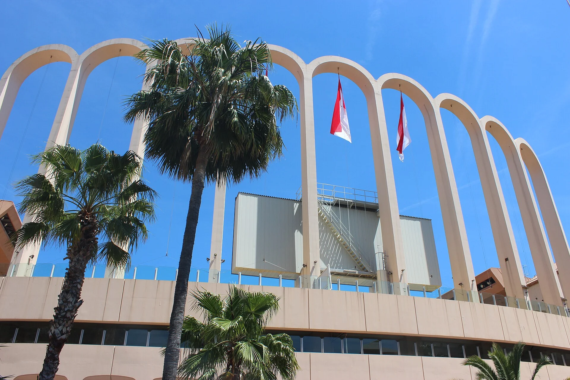 Tall pale arches of Stade Louis-II against a blue sky, with palms in the foreground.