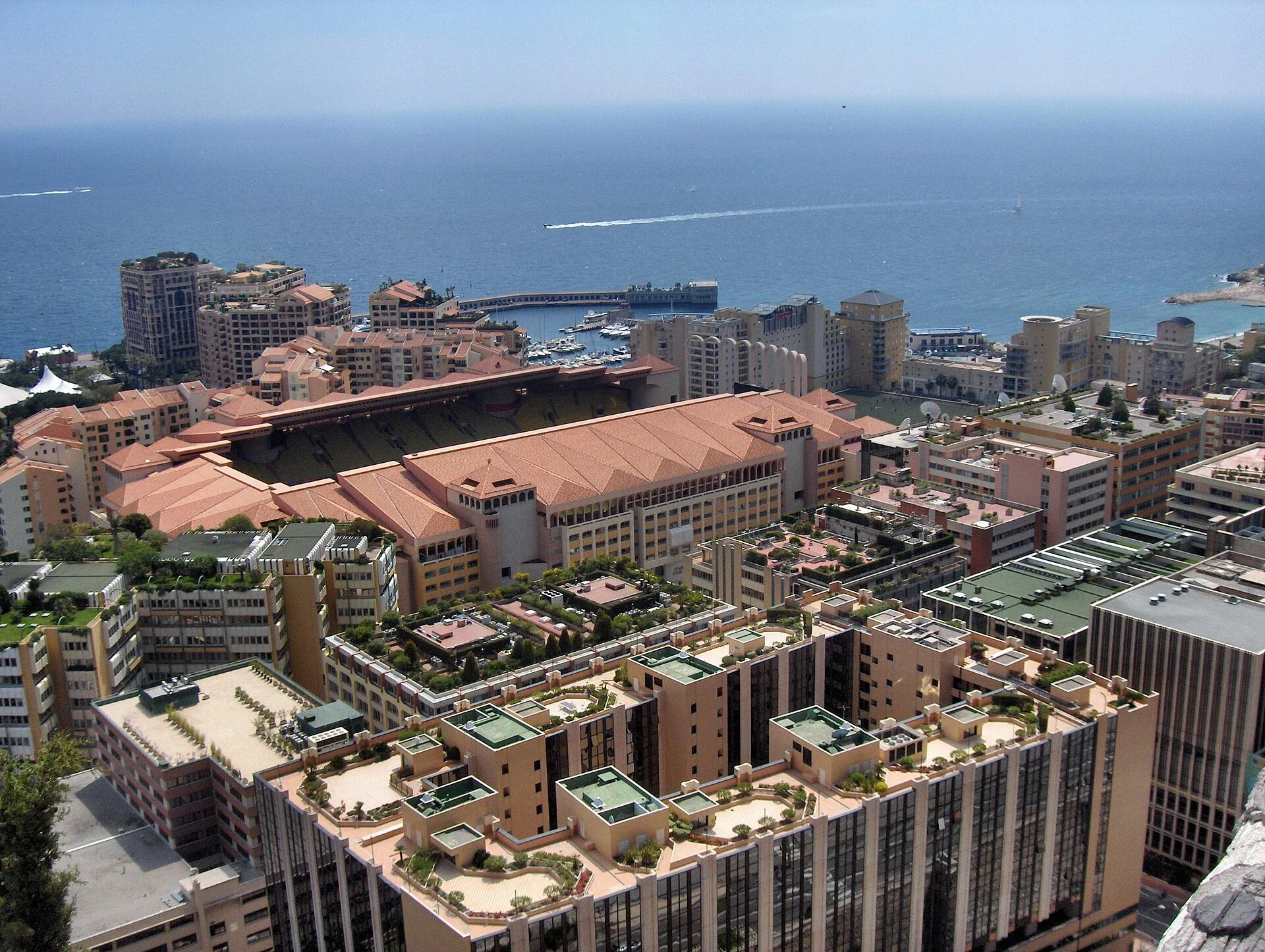 Stade Louis-II’s terracotta roof and athletics bowl amid the pale buildings of Fontvieille, with the Mediterranean Sea behind.