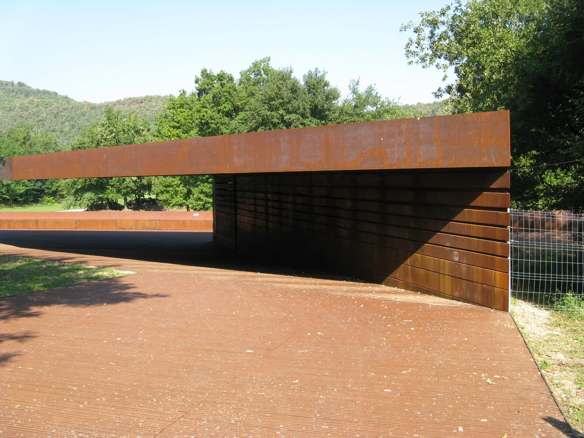 Rust-coloured entrance pavilion at the Tossols-Basil athletics ground beneath a clear sky and green trees.