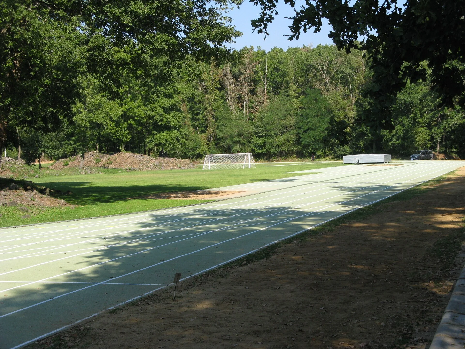 Pale green running lanes at Tossols-Basil bordered closely by dense woodland, with trees casting shadows across the straight.