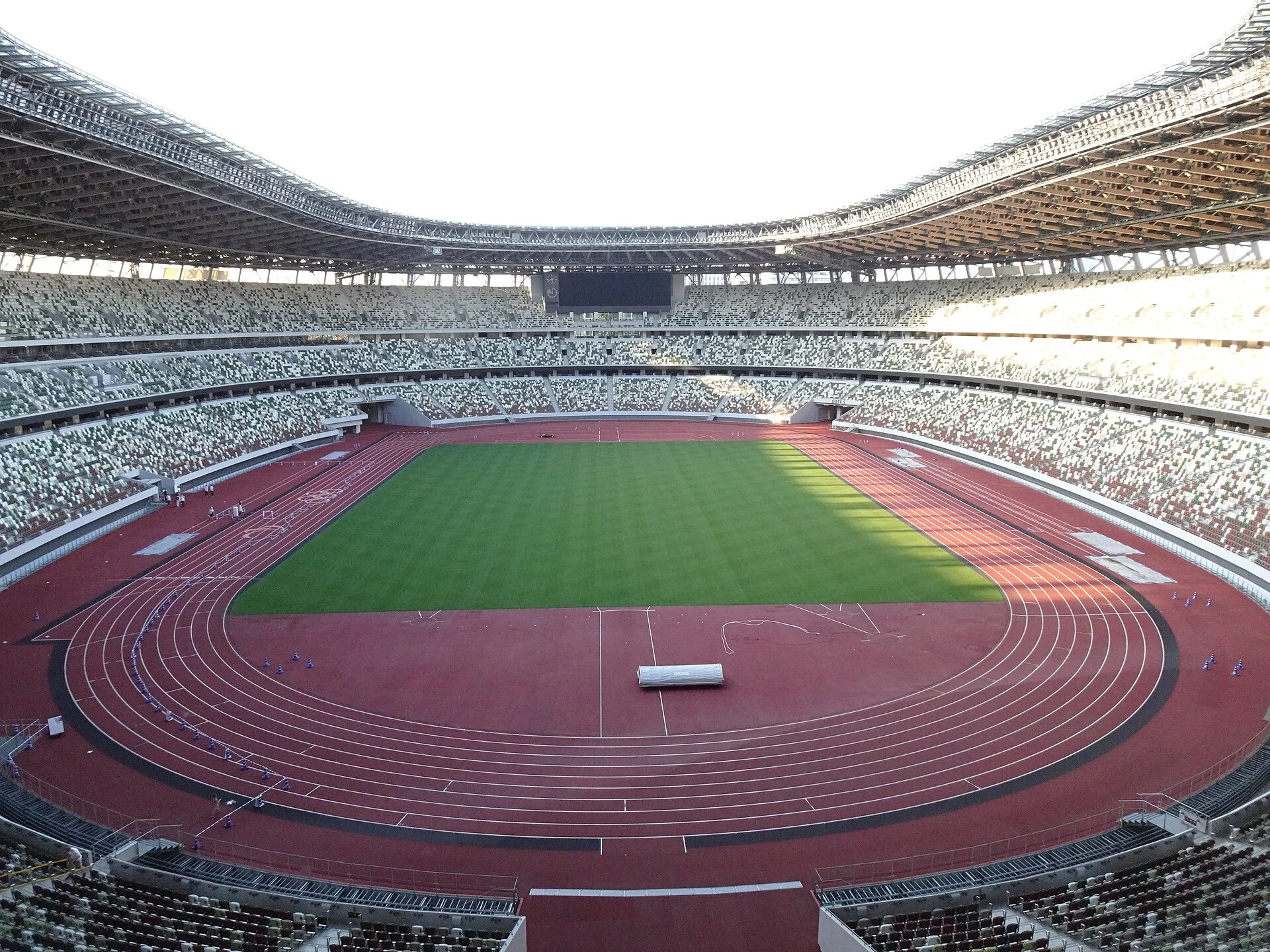 The red nine-lane running track and green field of Japan National Stadium seen from high in the stands, beneath a broad roof opening.