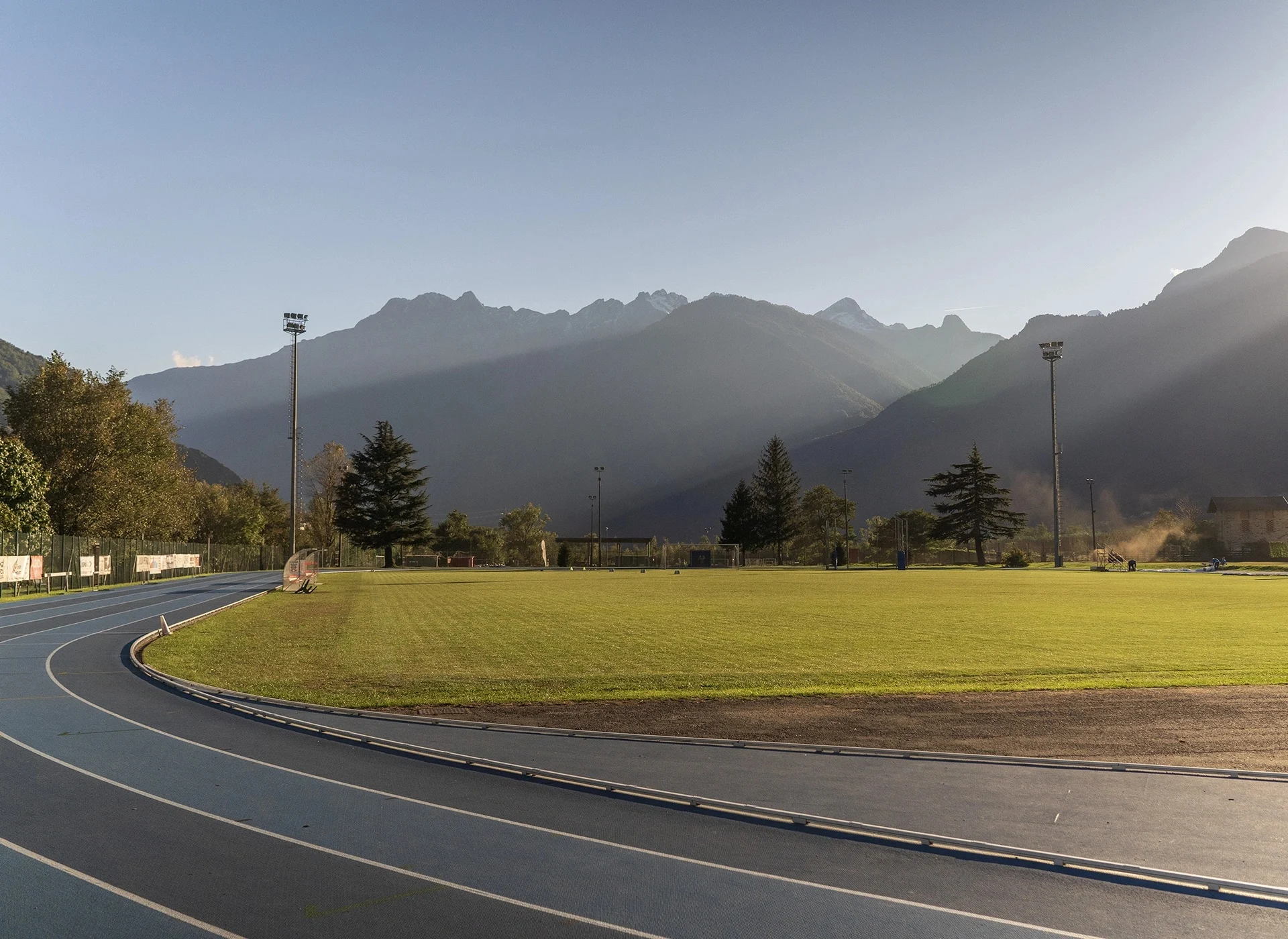 A blue-grey running track curves around a green infield beneath a layered mountain skyline in hazy sunlight.