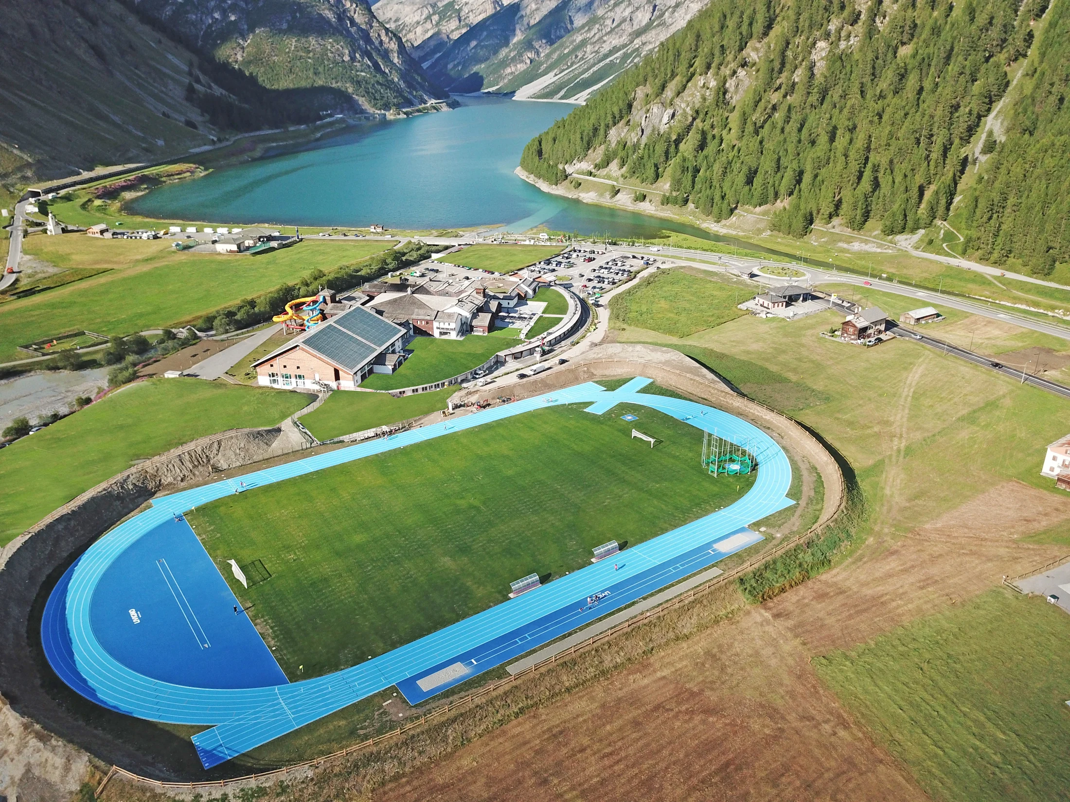 The blue running track at Livigno set beneath mountain slopes beside a turquoise lake.