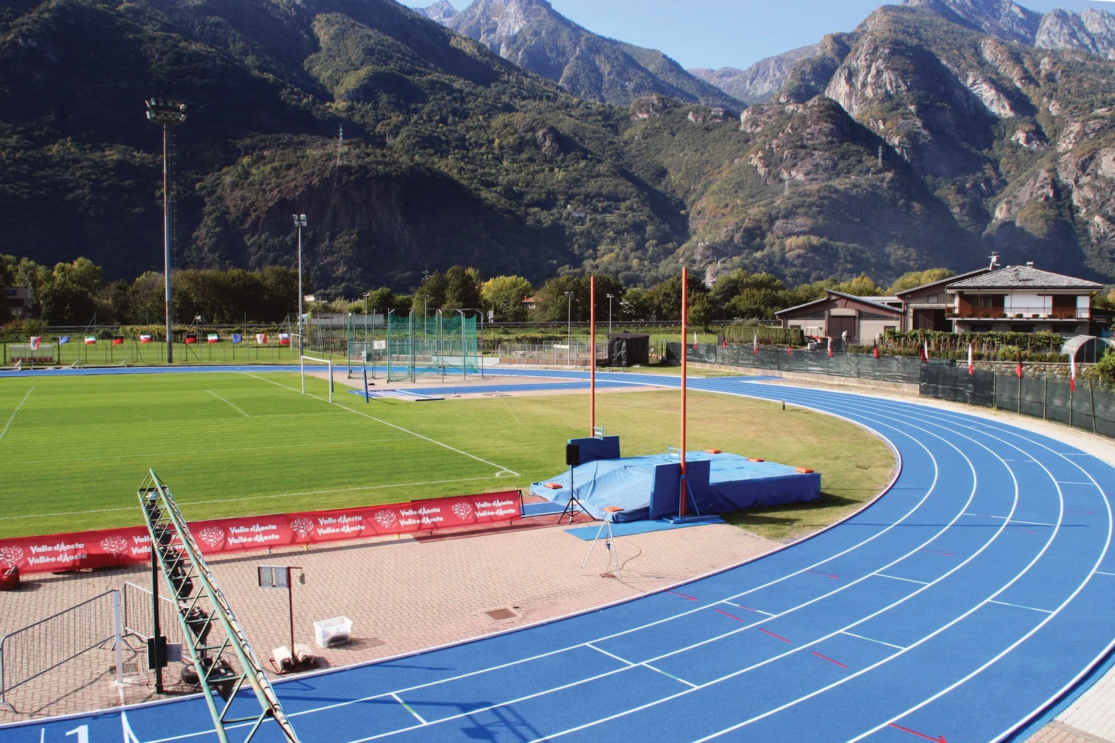 Blue running lanes sweep past a jumping area, with steep rocky mountain slopes immediately beyond the Donnas stadium.