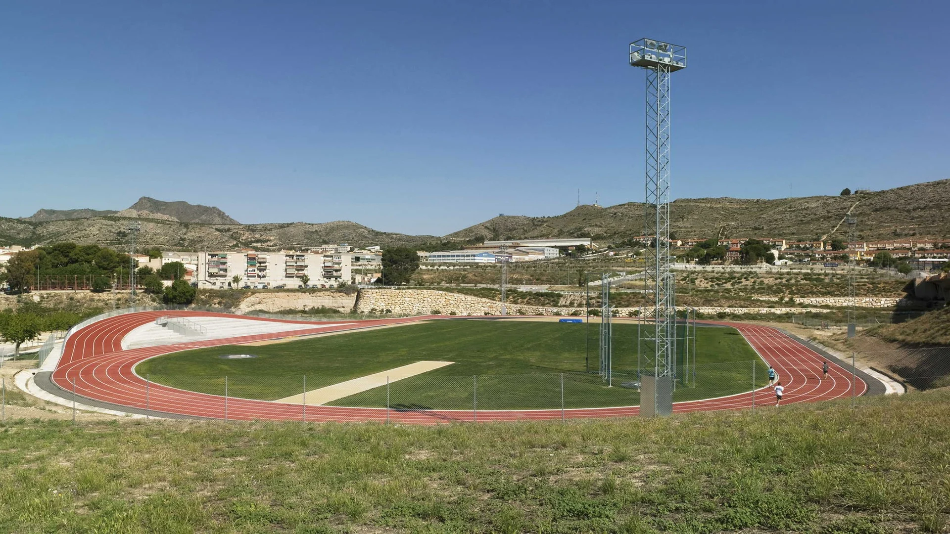 A red athletics oval with an additional undulating track section rising over a low white building, set among dry hills in Elda.