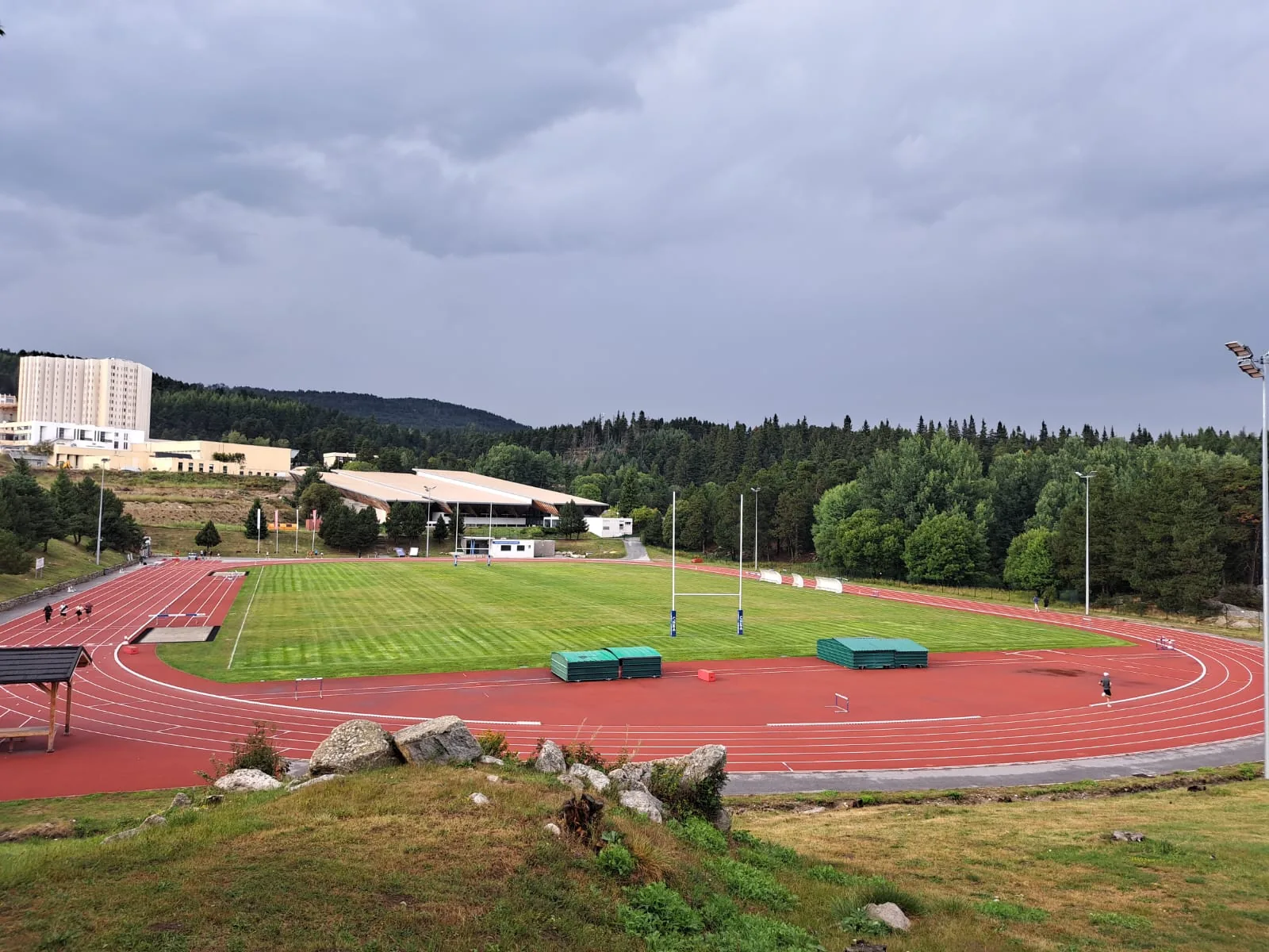 The red track and green infield of Stade Colette Besson at Font-Romeu, with the CNEA buildings, pine forest and a stormy sky beyond.