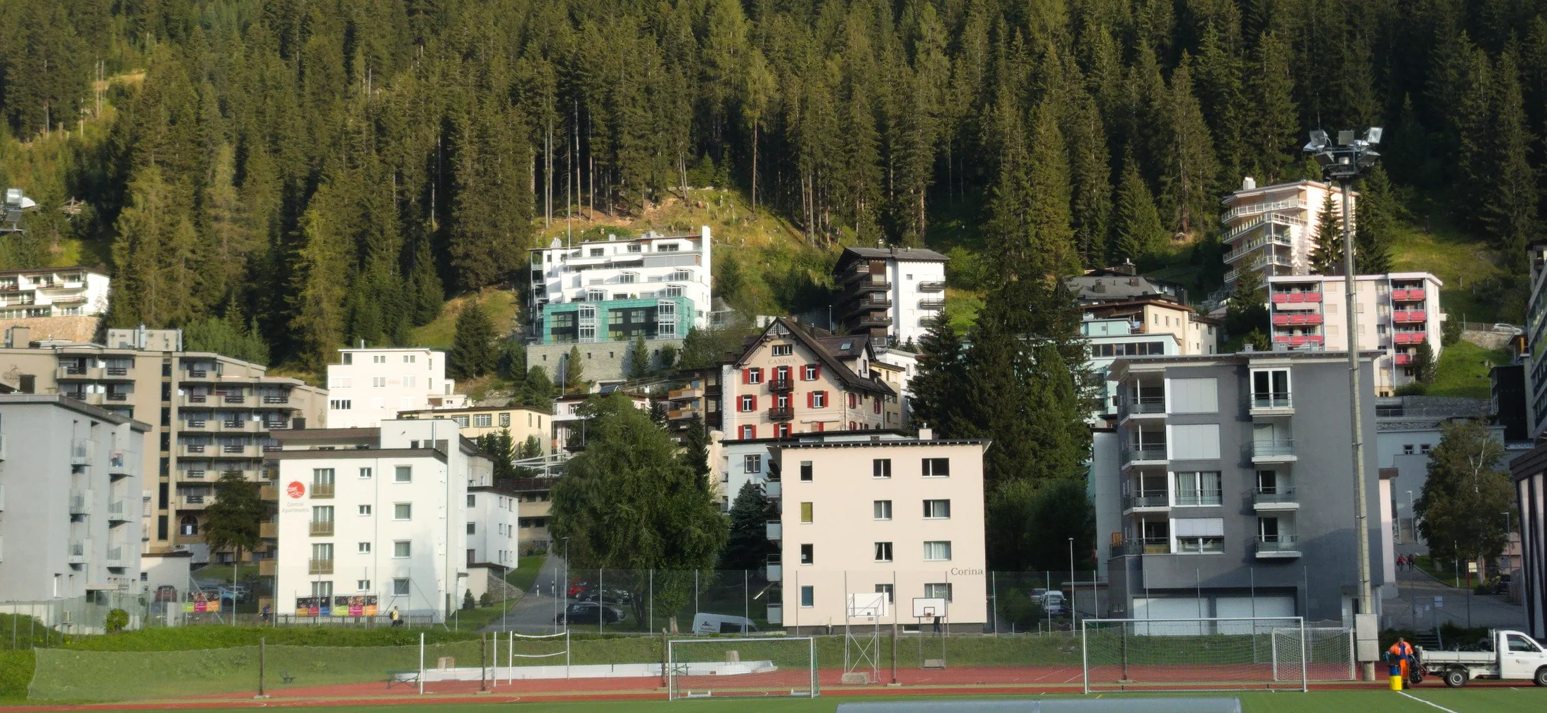 Red lanes and a pitch at the Davos Sportzentrum, with apartment buildings and steep pine forest rising behind.