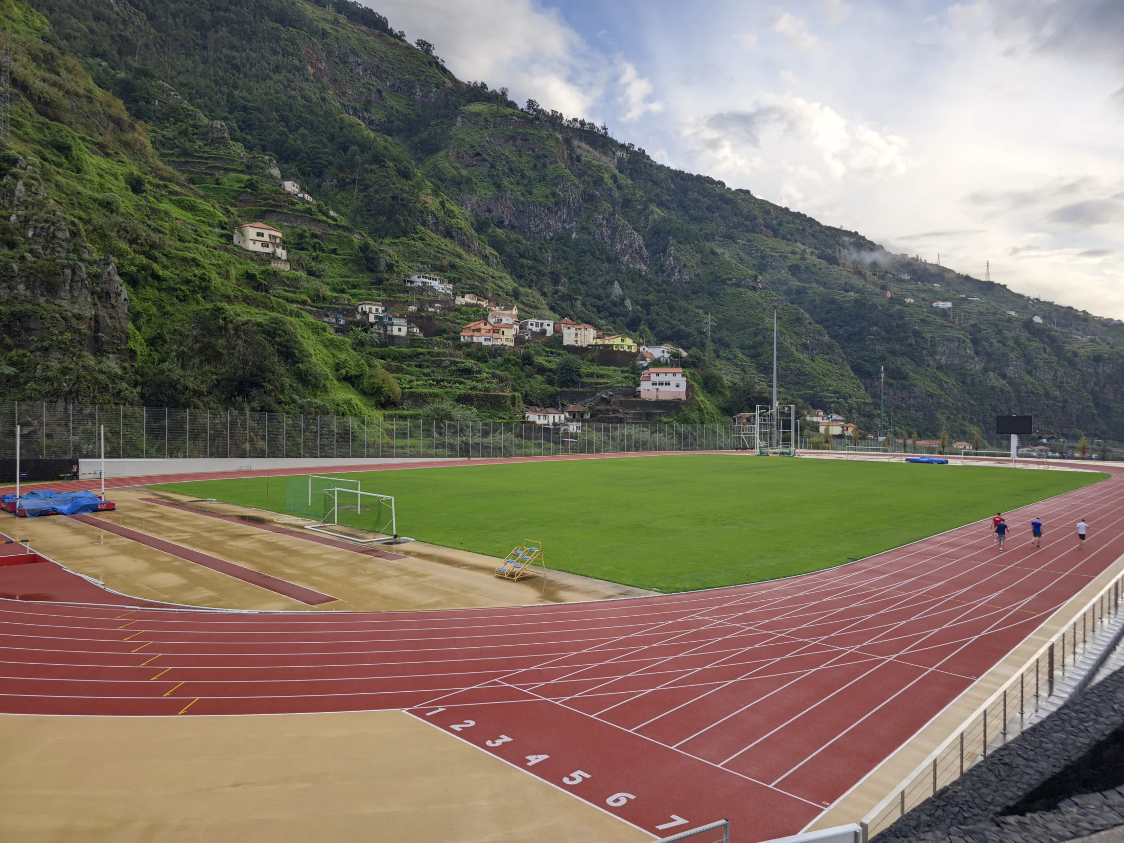 Numbered red lanes curve around a green pitch at the Centro Desportivo da Madeira, beneath steep terraced slopes dotted with houses.