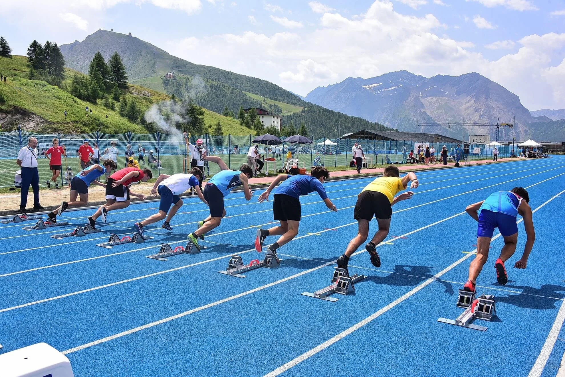 Sprinters leave their blocks on blue lanes with mountains and the sports-centre roof behind them.