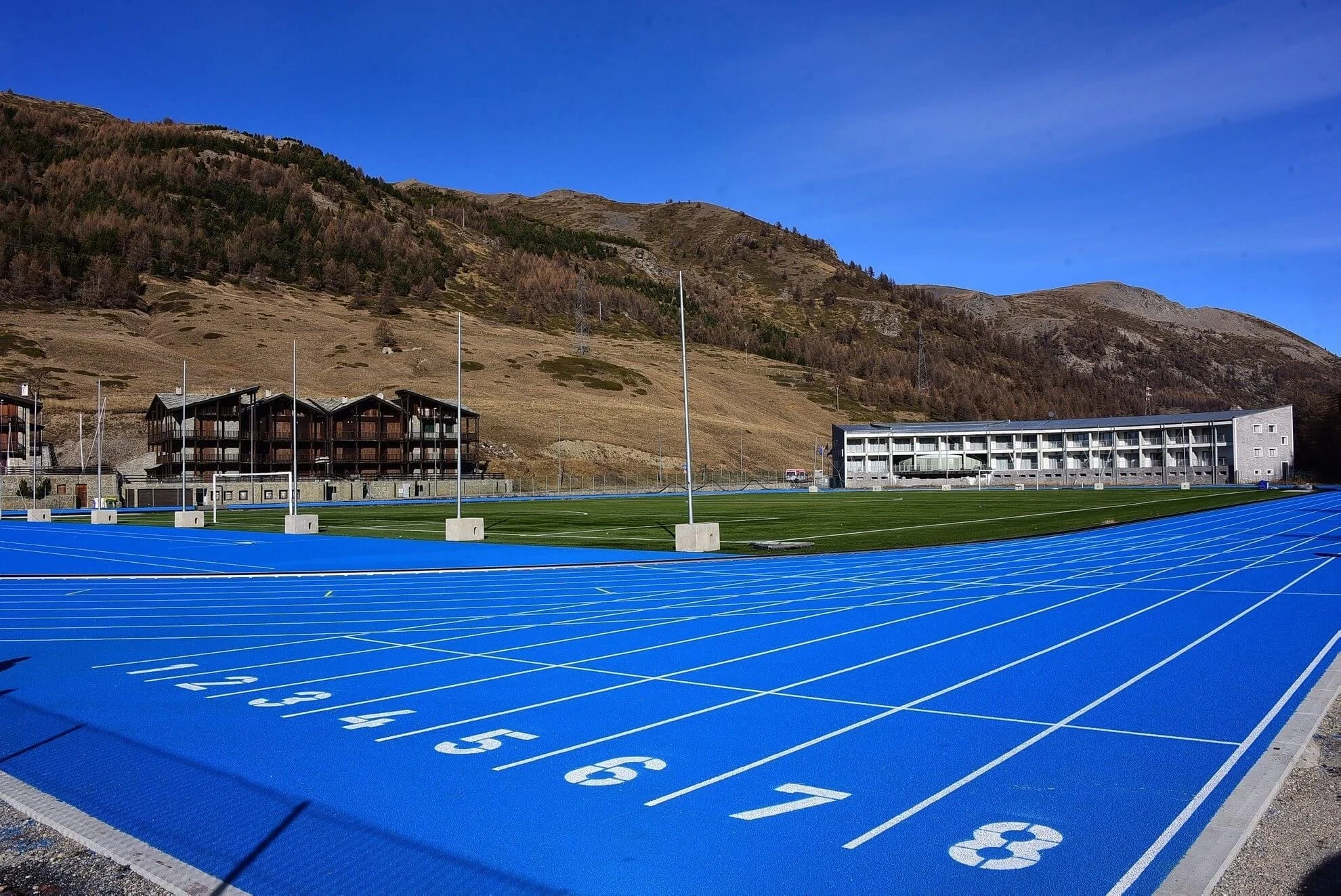 Eight vivid blue running lanes in a mountain basin beside a low sports-centre building, with Alpine slopes rising behind.