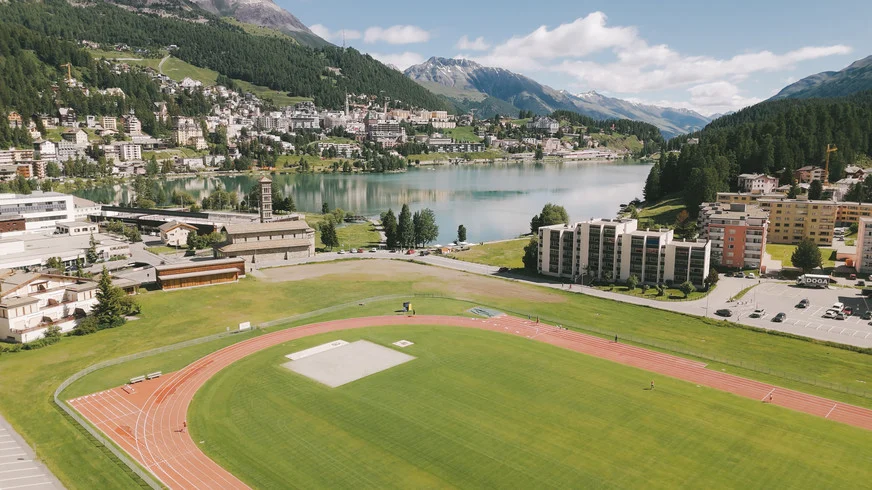 The red six-lane athletics track and green infield beside Lake St. Moritz, with forested slopes and Alpine mountains beyond.