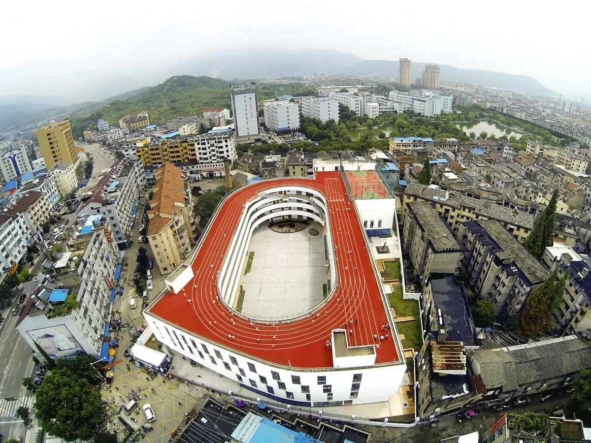 A high aerial view showing the complete red rooftop oval around the courtyard of Tiantai No. 2 Primary School, with streets and buildings below.