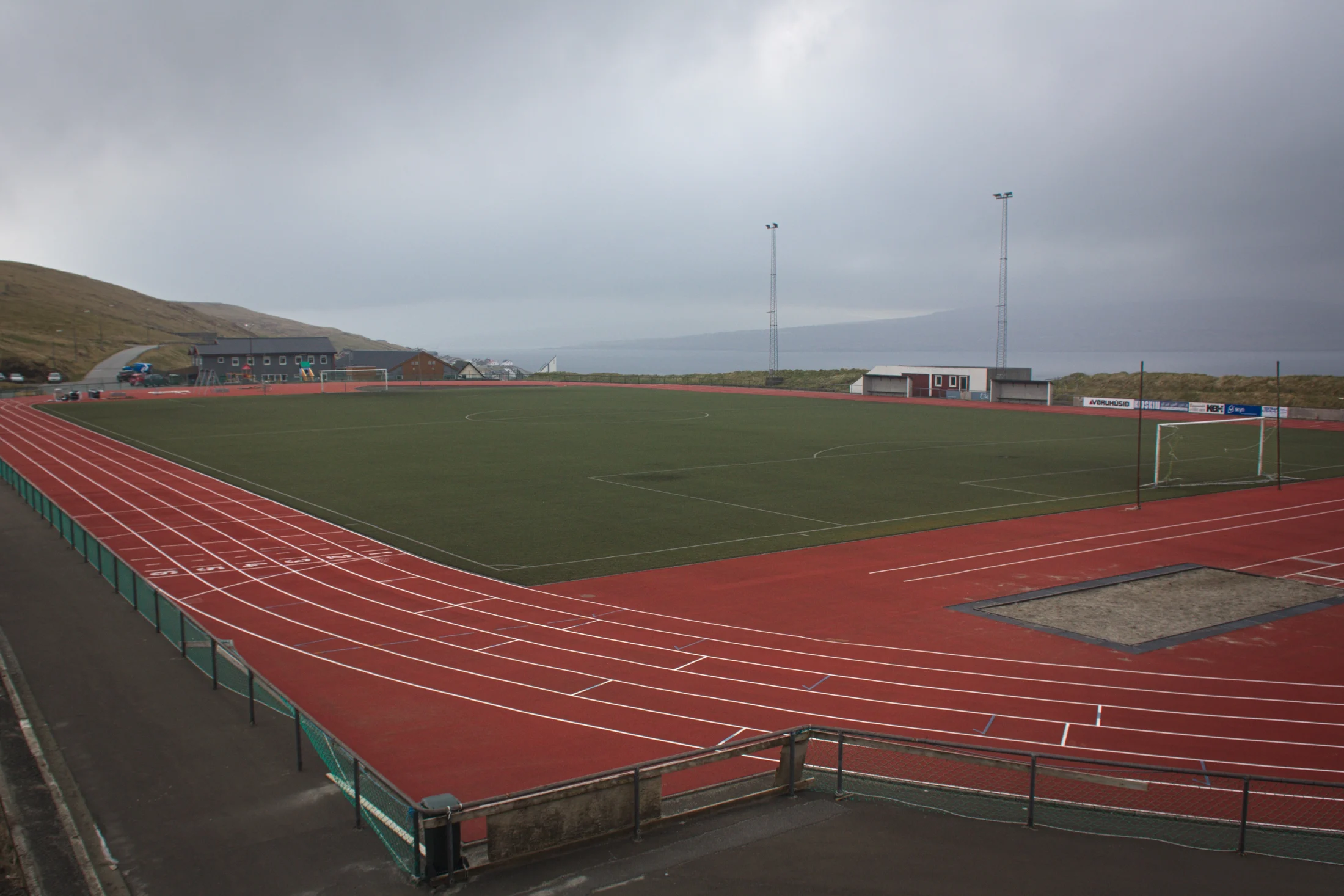 Red running lanes around the lower field at Svangaskarð, with low treeless hills and a misty horizon beneath grey cloud.
