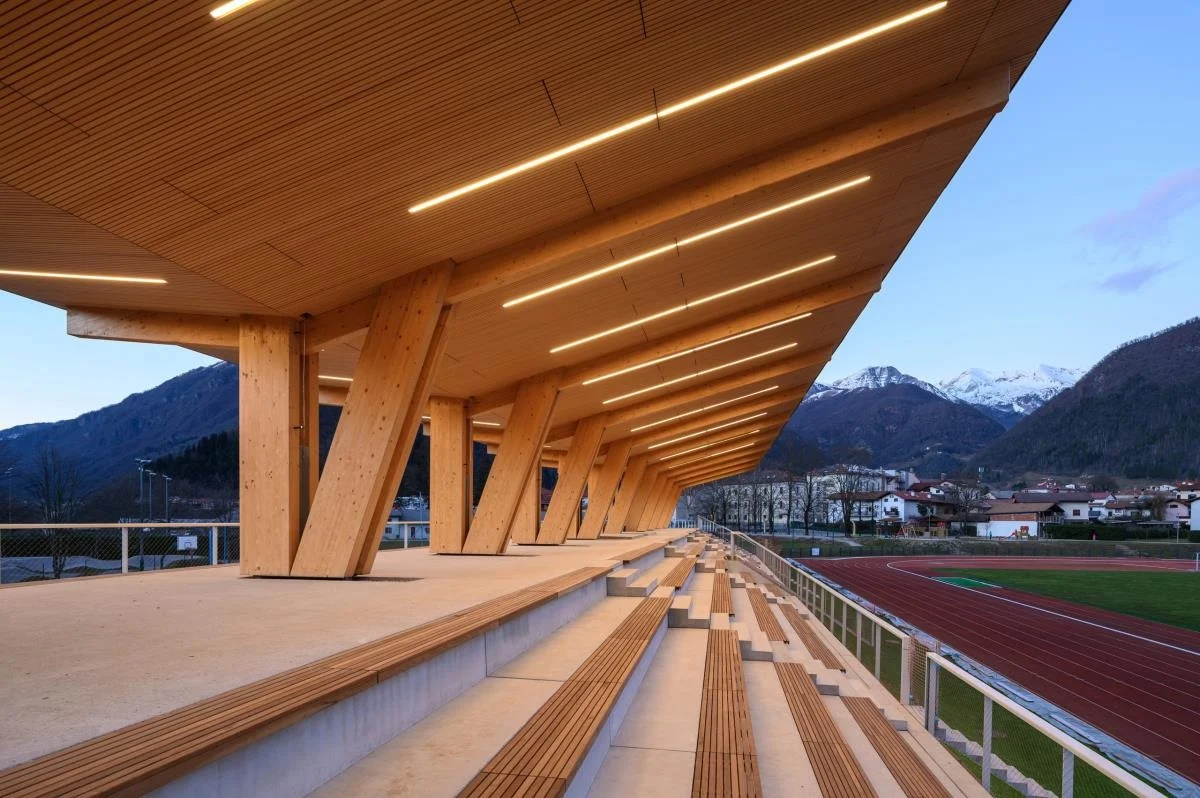 A long timber grandstand glowing beneath its roof at dusk, with the red Tolmin athletics track and snow-tipped mountains beyond.